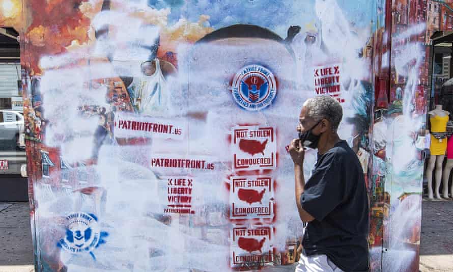 A Black man wearing a face mask walks past a mural of George Floyd that has been defaced with white paint and stenciled graffiti promoting white nationalist group Patriot Front. 
