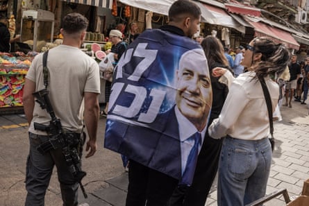 A man with a flag wrapped around his shoulders featuring an image of Israel’s prime minister, Benjamin Netanyahu, walks through Mahane Yehuda market in Jerusalem. On his left is a man carrying a semi-automatic weapon, on his right is a woman