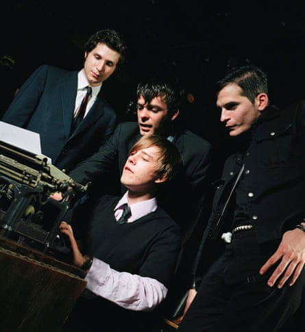 Four men weearing black, two with ties, sitting and standing around a typewriter