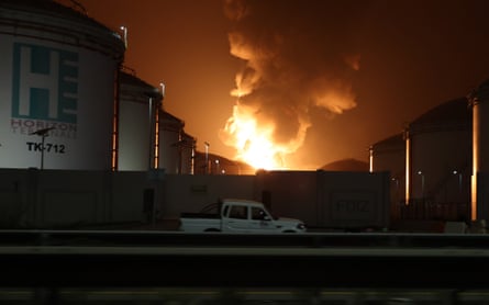 Flames rise at night over large oil tanks.