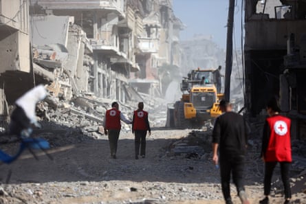 Three ICRC officials accompany a man in black clothing through a ruined street towards an excavator