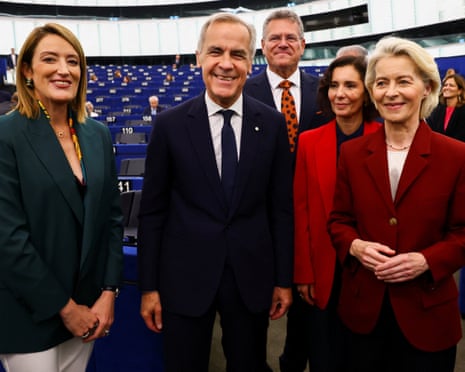 Metsola, Carney and von der Leyen stand in front of 2 other people in the parliamentary chamber. All are smiling