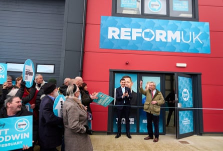 Nigel Farage and candidate Matt Goodwin stand ready to cut a ribbon at the doorway of the building, which has blue Reform UK signage on a red facade. People are standing nearby holding blue Reform placards.