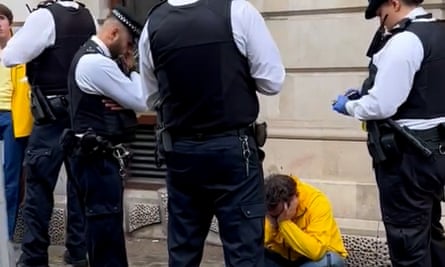اسمیت با یک ژاکت زرد، نشسته روی بند خود و سرش در دستانش، در محاصره افسران پلیس