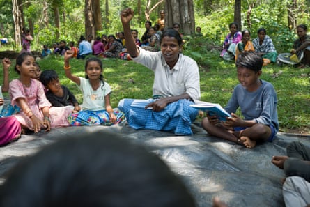 A young Indian man in a lungi sits in a forest clearing in a circle of children