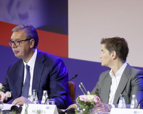 Ana Brnabić and Aleksandar Vučić at a formal meeting. She is 50 with very short brown hair and wears a grey jacket; she is turned towards Vučić. He is in his mid-50s and has short grey hair with a side parting. He is very tall, and wears glasses. qhxidiqxkiqxkinv