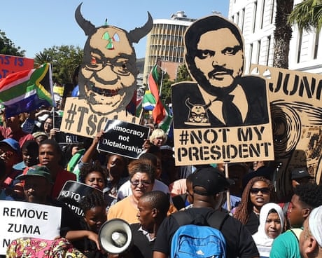 A crowded street protest with placards of an African man caricatured with horns (Zuma) and another with an Indian holding the horned Zuma in his pocket. Slogans include phrases such as 'Not my president' and 'remove Zuma'