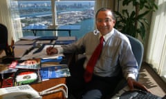 Man wearing glasses, buttondown and red tie sits at desk with a waterfront view in the behind him