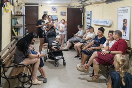 Mothers with young children and older people sit on benches or stand by prams in a small underground room