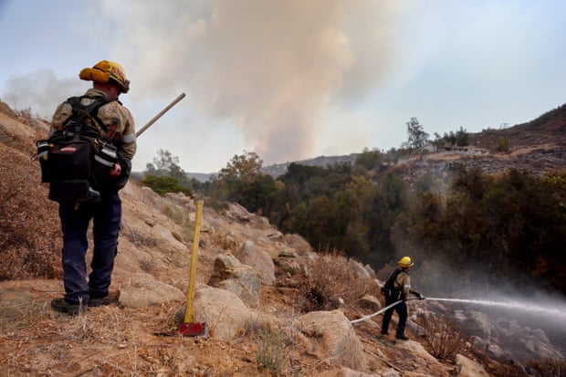 Two firefighters stand on a ridge aiming a fire hose at a hot spot as a plume of smoke rises in the background.
