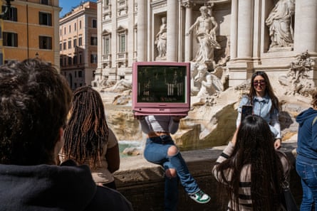 one young woman holds a computer screen in front of her head while another sits to have her picture taken.