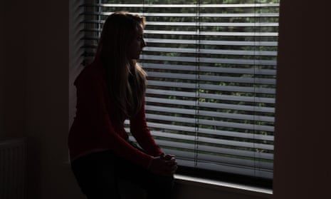 Young female sitting alone in a dark room looking out through a window blind.