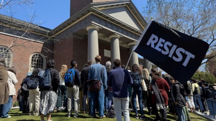 People gather outside a building, one carrying a large flag that bears the word 'Resist'