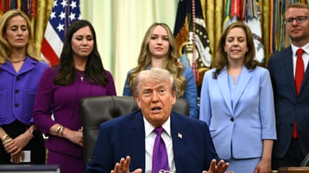 Donald Trump sitting at a desk in the Oval Office with people standing behind him