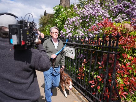 A cameraman to his left, Peter Mandelson walks his dog by some shrubbery