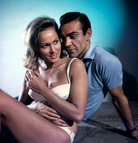 Andress sits with Connery; he has his chin on her shoulder. She is wearing a white bikini and he wears a blue polo shirt. qhiukiuiqkqinv