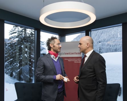 Two men speak face-to-face in a room. Snowy mountain scenes can be seen through the windows.