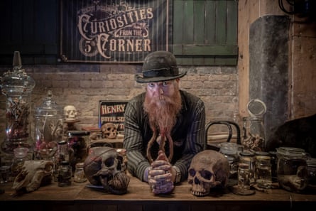 Henry Scragg sits at a table bearing skulls, and jars filled with curios