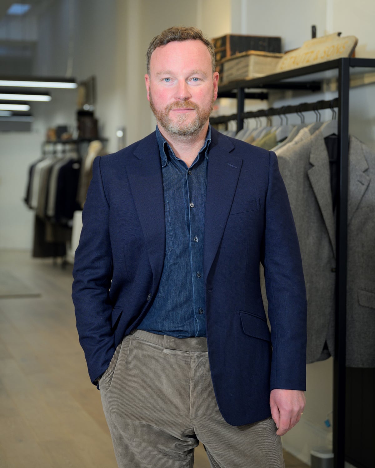James Sleater in suit jacket and chinos, standing in fronts of suits on rails