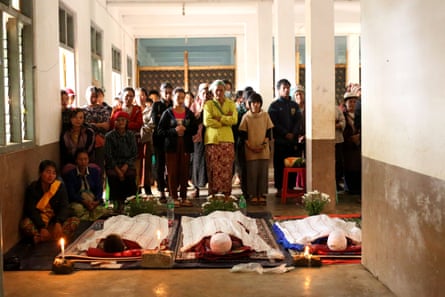 People stand and pray next to a line of three bodies laid out on the ground
