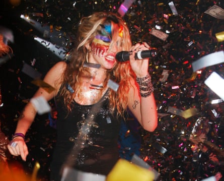 A blond woman in a low cut black dress with loads of face paint grabbing a pic - with a lot going on around her