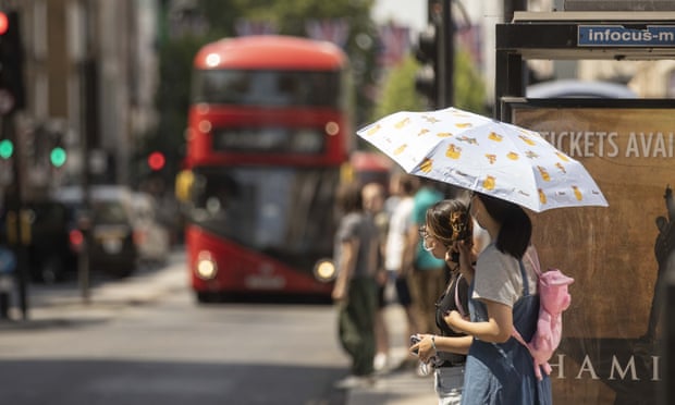 Women holding an umbrella to protect themselves from the sun wait at a bus stop in London