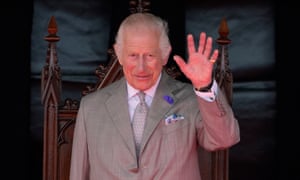 King Charles in grey suit waves in midshot with ornate wooden chair behind