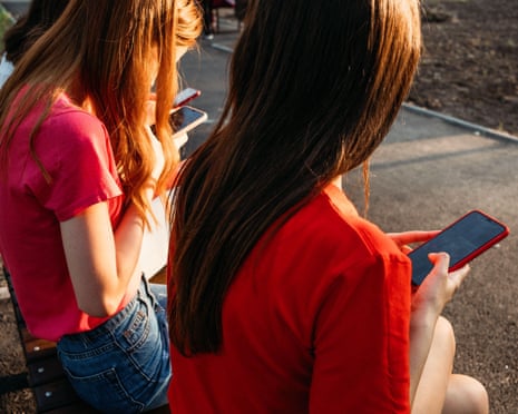 Girls sit on a bench, both looking at their mobile phones