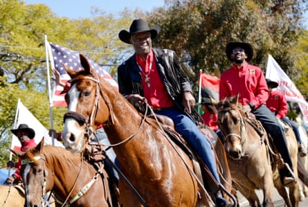 Older middle-aged Black man, wearing black cowboy hat, red collared shirt, black leather jacket and jeans on brown horse, followed by other Black men in red collared shirts and black cowboy hats, some holding American flags and one holding the pan-African flag.