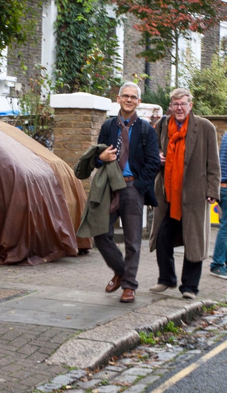 Alan Bennett dressed in a big brown coat, red/orange scarf, black trousers and brown shoes, walking alongside Rupert Thomas, dressed in a navy blazer, mid-blue shirt, spotty tie and loosely tied patterned silk scarf, brown belt and shoes holding a grey coat over his right arm, walking along a smart residential street in London, both slightly smiling at the camera
