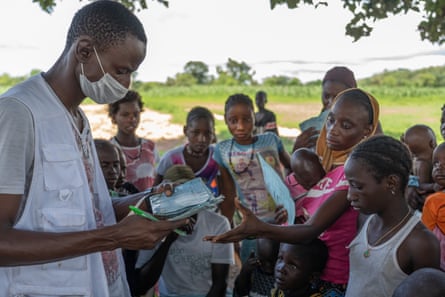 A man wearing a white vest and a facemask holds paperwork and a pen. A woman standing in front of him reaches out to take her documents back