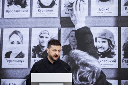 Volodymyr Zelenskyy speaks at a lectern, with a backdrop showing photographs of people and their names in Cyrillic