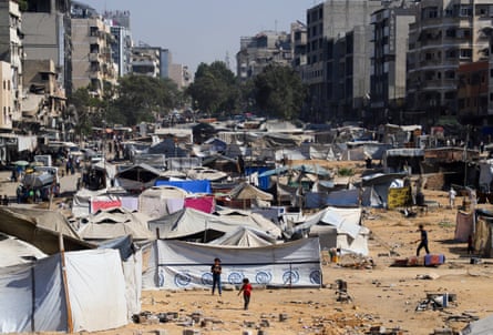 makeshift tents and shelters are set up on sand and rubble in a space between damaged buildings in a street which looks as if it may have previously been a wide road or boulevard. Children are playing on the sand.