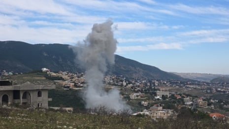 Grey smoke rises into a blue sky, with buildings and mountains in the background.