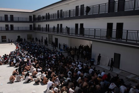 A large number of people sit on the floor outside in what appears to be a detention complex