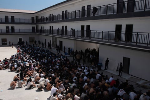 A large number of people sit on the floor outside in what appears to be a detention complex