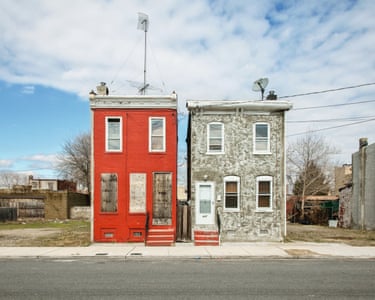 a red-painted three-storey brick house, its ground floor boarded up, stands next to an occupied mottley grey one. Empty lots either side
