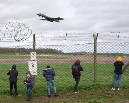 People watch and take pictures as a military plane takes off in an airfield.
