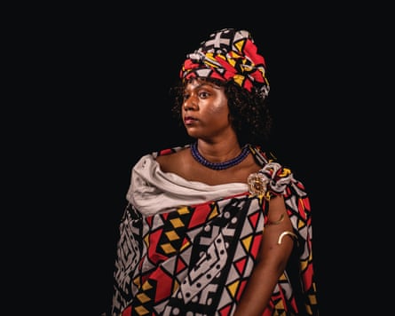 A woman in an African print cloth and matching headdress, wearing beads and an armlet, looks off to one side in a statuesque pose