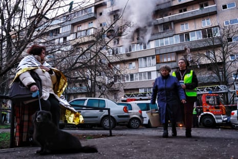 Psychologist leads an elderly woman while smoke rises from damaged residential building after Russian drone-and-missile attack on Kyiv, Ukraine.