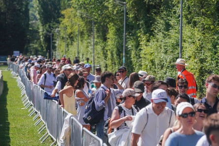 People queuing in the sunshine, with trees on one side and a fence on the other