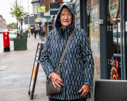Delphine Hazeldon, in a hooded leaf-print coat with a crossbody bag, smiles on a shop-lined street