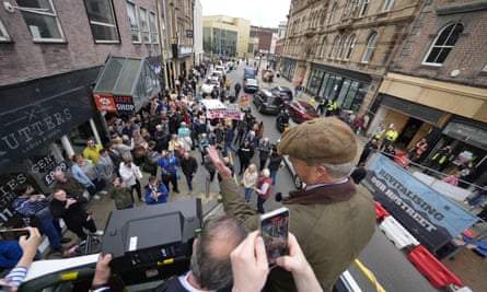 Back view of Nigel Farage on top of open bus with demonstrators with placards below