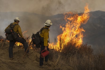 Firefighters look down a hillside as a fire burns in the brush in front of them.