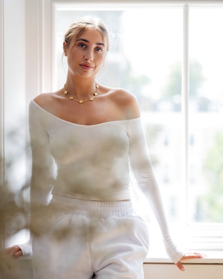Phillips poses for a bright portrait against a window, wearing white jogging bottoms and an off-the-shoulder white top