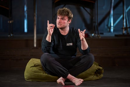 A young man dressed in a black sweatshirt and tracksuit trousers with light brown hair sits with his legs crossed on a green cushion and gestures with his hands as he speaks.