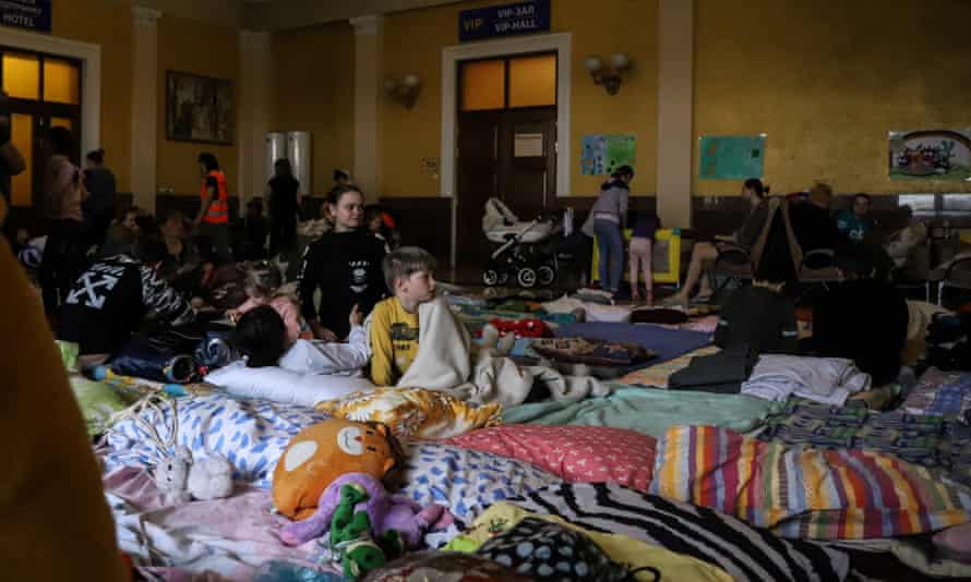 Camp beds, covered in brightly colored duvets and quilts, are crammed together in a large room.  Three children in the foreground