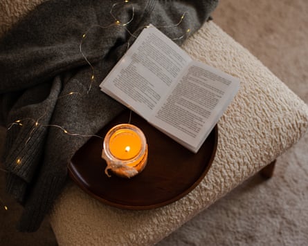 Scented burning candle with an open paper book on a wooden tray and a knitted textile on a chair