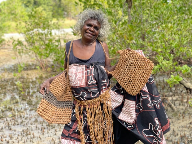 Esther Yarllarlla, standing by Djomi with nja-djéngka (dilly bags), mókko pubic covering, and fabric design, 2022.