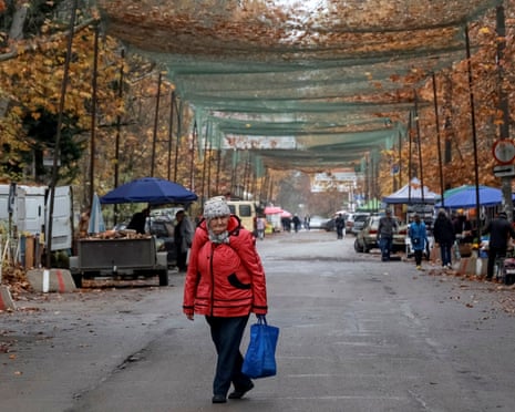 An old woman with a shopping bag crosses a road lined with market stalls with netting draped over it